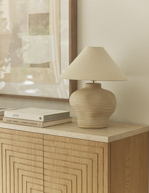 The Wrinkle Table Lamp by Sarah Sherman Samuel in beige ceramic with a matching shade rests on a light wood cabinet, accompanied by stacked books and framed abstract art in a gently lit room.
