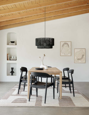 The Abode geometric two-toned kand-knotted floor rug by Élan Byrd with woven border sits in a dining room with black ida dining chairs, a black jute chandelier, and a sloped wooden beamed ceiling