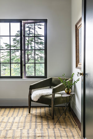 The Lyssa olive velvet accent chair sits in the corner of a room with a striped rug and a white sheepskin blanket thrown atop it.