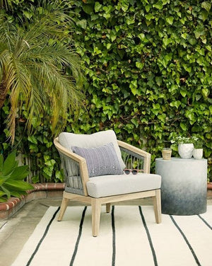 The Pira gray and white ombre indoor and outdoor side table sits in an outdoor space with a gray linen accent chair, a black striped rug, and an ivy wall