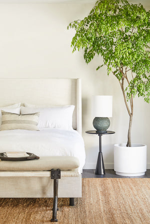 The Arashi black metal side table with rounded top and sculptural base sits in a bedroom with a talc linen adara bed, a lamp with a rounded stone base, and a jute rug