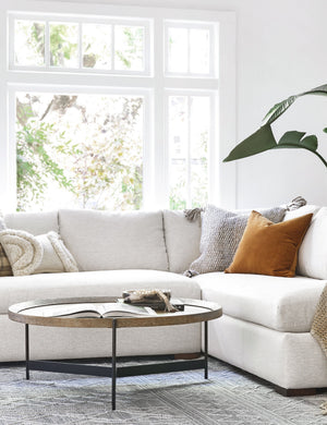 The Becca coffee table with rimmed oak top and iron tripod-style base sits in a living room with a linen sofa and a large white framed window in the background