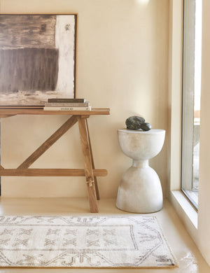 The Arlene craftsman-style antiqued teak wood console table has a neutral abstract painting leaning on top of it with a stack of books, and a light gray antiqued stone rounded side table next to it.