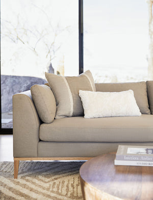 The Samaire shearling white plush lumbar pillow sits in a living room atop a gray linen couch in front of a brown and gray patterned throw pillow