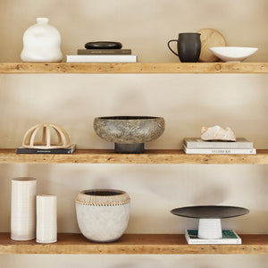 The Lakshmi terracotta bowl with antiqued black finish and woven base sits on a wooden shelf with other centerpiece decor and stacks of books