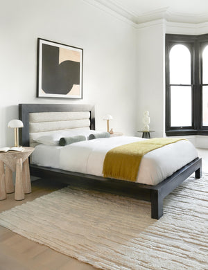 The Lelani table lamp with half-moon shade on a gold base sits in a bedroom atop a sculptural black nightstand to the left of a bed with a wooden framed boucle headboard