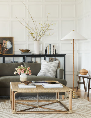 The Zora floor lamp with polished base and brass knob detailing sits in a living room with white accented walls, a sage velvet sofa, and a woven coffee table