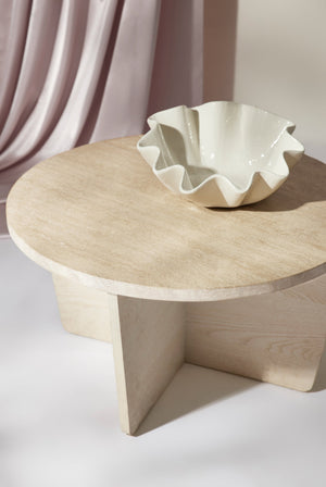 The Ruffle ceramic cream centerpiece bowl by Regina andrew sits atop a white wooden coffee table with a dusty pink curtain draped in the background