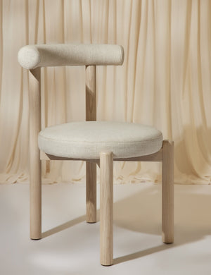 Angled left view of the Dame minimalist rounded open-back dining chair with ivory upholstered seat and ash wood frame on a tan fabric draped background