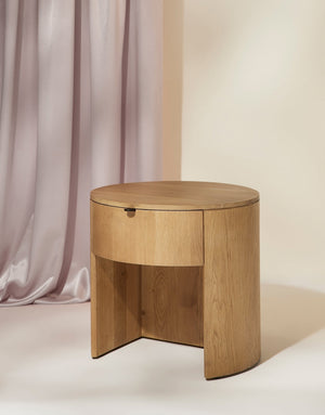 Angled right view of the Kono round solid oak nightstand with one drawer on a purple draped backdrop