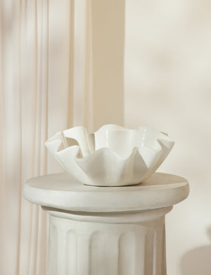 The Ruffle ceramic cream centerpiece bowl by Regina andrew sits atop a white pedestal with a white curtain draped in the background