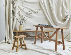 The Arlene craftsman-style antiqued teak wood console table, along with the matching Arlene stool and bench are displayed against a wall draped in light tan fabric.