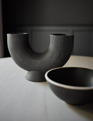 The Damien half-circle sculpture vase by Ateriors with matte black glaze finish sits atop a white wood table with a ceramic bowl to the right of it