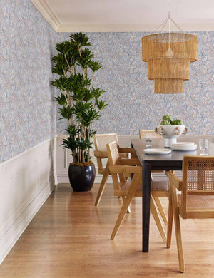 The Chavette three-tiered jute-wrapped chandelier hangs over a black dining table surrounded by light wood chairs with cane backs in a dininr room with blue abstract wallpaper.