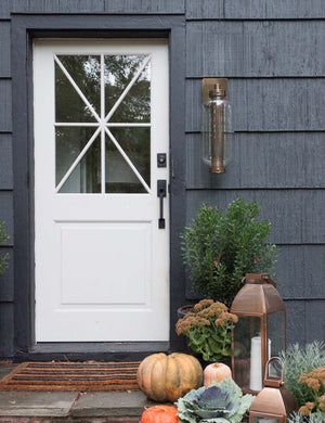 The Romelia indoor and outdoor sconce with metal mesh encased bulb and brass hardware hangs out the exterior of a black paneled home to the right of a white door