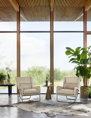 Two Dove ivory leather accent chairs sit in a living room with floor to ceiling windows and a metal side table in between them.