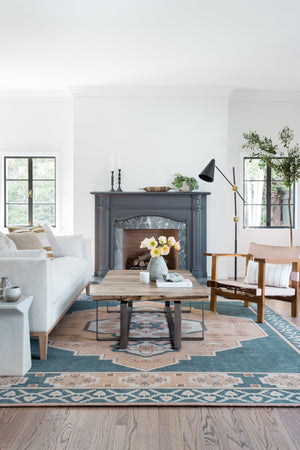 The Tilda t-shaped marble side table sits in a living room with a wooden slab coffee table, ivory linen sofa, and a large accented black marble fireplace in the center of the room