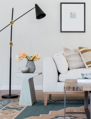 The Tilda t-shaped marble side table sits in a living room in between a golden and black floor lamp and an ivory linen sofa, all sitting atop a teal and light orange geometric rug