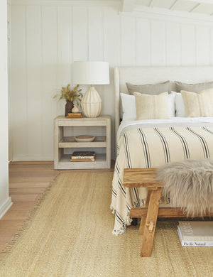 The Monte viso natural and black striped bed cover with fringed ends by House No. 23 sits at the end of a linen adara bed in a bedroom with a jute rug and a white wooden nightstand
