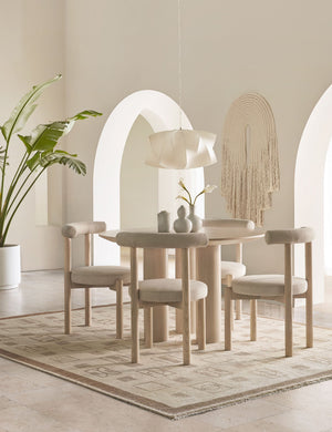 Four Dame minimalist rounded open-back dining chairs with ivory upholstered seat and ash wood frame surround a round light wood dining room with a sculptural white pendant light above and white and tan geometric print rug below.