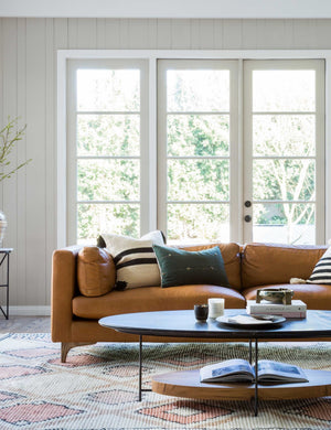 The Jocelyn brown leather sofa sits in a living room in front of white-framed french doors, behind a black oval coffee table, sitting atop a diamond patterned rug.
