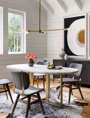 The Kara oval dining table with white marble top and brass base sits in a dining room with an animal skin rug, black woven dining chairs, and white paneled walls