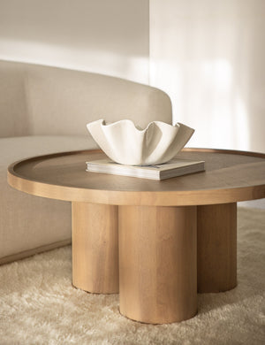 The Ruffle ceramic cream centerpiece bowl by Regina andrew sits in a living room with a circular wooden coffee table, plush carpet, and white linen sculptural sofa