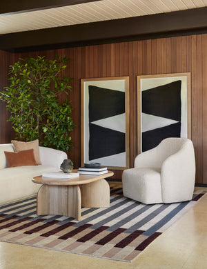 The Ada solid oak oval coffee table sits in between a curved ivory boucle couch and accent chair in a wood paneled room with two large vertical black and white geometric paintings and a graphic neutral striped rug.