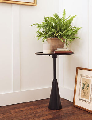 The Arashi black metal side table with rounded top and sculptural base sits in the corner of the room with white accented walls, a terracotta planter, and a floral wall art