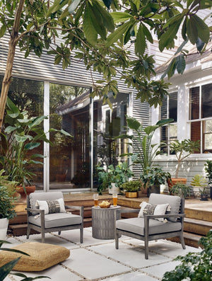 The Glyn concrete indoor and outdoor side table with carved finish sits in an outdoor space between two gray wooden framed accent chairs atop a tiled concrete floor